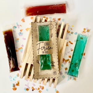 May include: A top-down view shows several brightly coloured ice pops in clear plastic packaging, arranged with a wooden beach chair and confetti. A brown card with the text "COOLE FERIEN FÜR DICH" is in the centre. The pops are various colours.