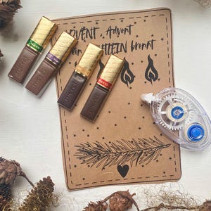 May include: Four chocolate bars with gold foil wrappers are arranged near a brown paper card with text and illustrations. The card has the words "ADVENT, Advent, Kerzelein brennt" and a drawing of a pine branch and a heart. A clear tape runner is also visible.