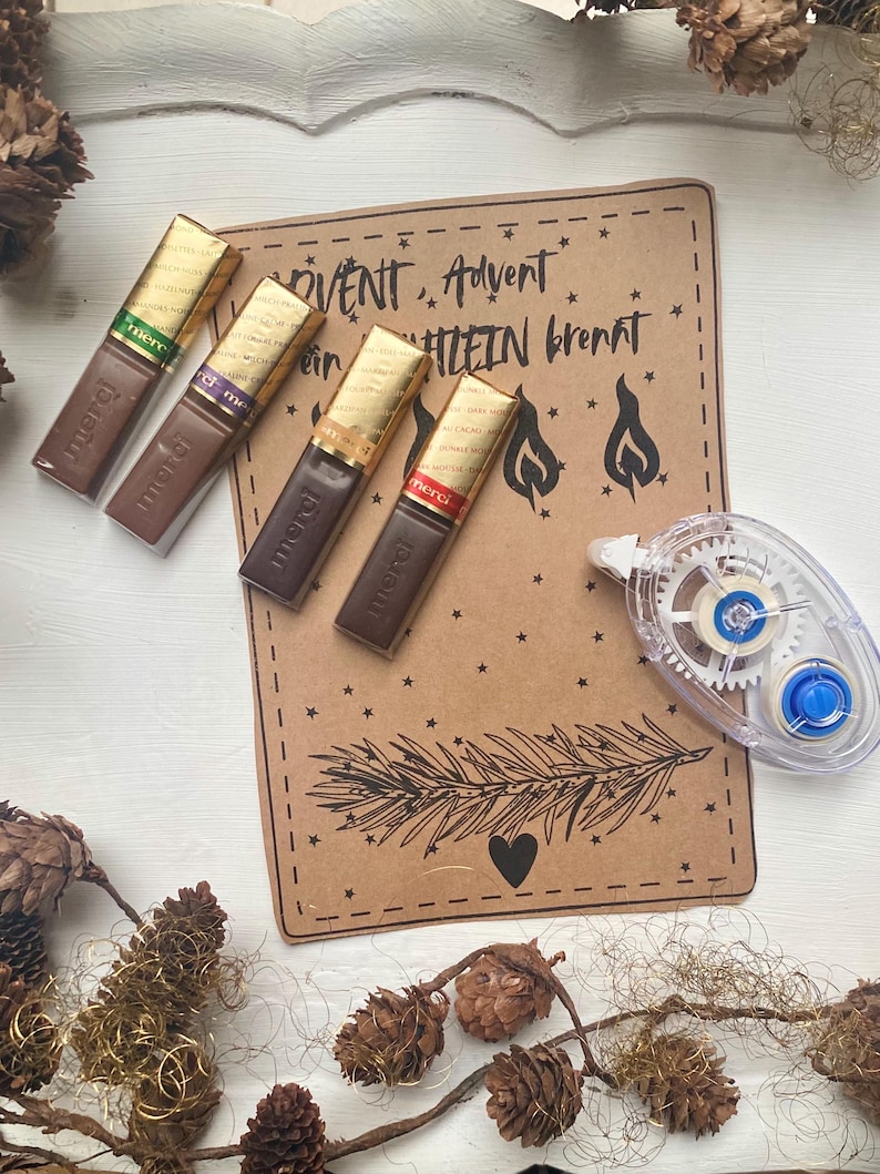 May include: Four chocolate bars with gold foil wrappers are arranged near a brown paper card with text and illustrations. The card has the words "ADVENT, Advent, Kerzelein brennt" and a drawing of a pine branch and a heart. A clear tape runner is also visible.