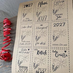 May include: A sheet of light brown paper featuring nine rectangular gift tags with dashed borders. Each tag displays black ink illustrations of fireworks and German text including 2027, Ahoi, Viel Gl&uuml;ck, and Silvester. A red curling ribbon lies to the left.