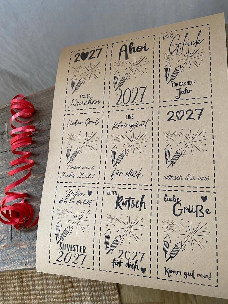 May include: A sheet of light brown paper featuring nine rectangular gift tags with dashed borders. Each tag displays black ink illustrations of fireworks and German text including 2027, Ahoi, Viel Gl&uuml;ck, and Silvester. A red curling ribbon lies to the left.
