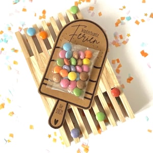 May include: A wooden popsicle-shaped card with the text "kunterbunte Ferien" and "für DICH" with a heart icon. A clear plastic bag containing multi-coloured candy-coated chocolate drops is attached to the centre of the card, which rests on a wooden rack.
