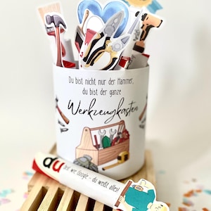Könnte beinhalten: Ein weißer zylindrischer Behälter mit Werkzeugkasten- und Werkzeugdesign, mit dem Text "Du bist nicht nur der Hammer, du bist der ganze Werkzeugkasten." Enthält Papierschnitte von Werkzeugen und ein "Best Dad"-Abzeichen.