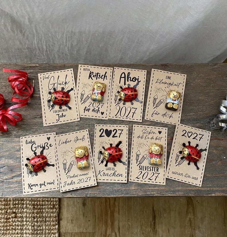 May include: Nine rectangular kraft paper cards featuring printed New Year greetings and illustrations of fireworks. Each card is adorned with a small chocolate figure, either a red ladybug or a gold bear, attached to the center of the card.