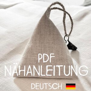 Op de afbeelding: Een beige linnen driehoekige tas met een koordsluiting en een zwarte clip. De tas heeft de tekst "PDF NÄHANLEITUNG DEUTSCH" erop gedrukt, met een Duitse vlag.