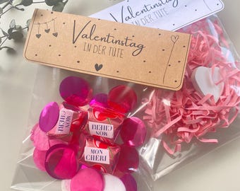 May include: Two clear cellophane bags filled with pink confetti, candies, and shredded paper. One bag has a brown tag that reads "Valentinstag IN DER TUTE" with heart illustrations. The other bag has a white tag with the same text.