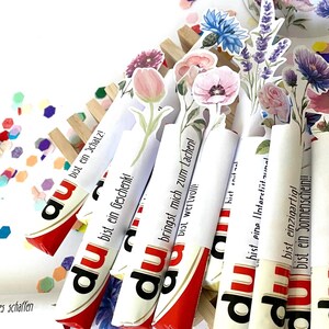 Pu&ograve; includere: Barrette di cioccolato Kinder Bueno con decorazioni floreali e testo in tedesco. Le barrette sono disposte in un supporto di legno e circondate da coriandoli. Confezione rossa e bianca.