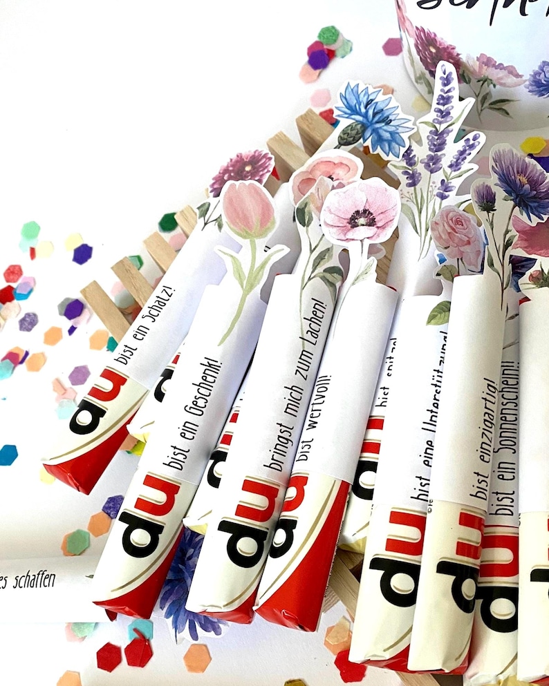Pu&ograve; includere: Barrette di cioccolato Kinder Bueno con decorazioni floreali e testo in tedesco. Le barrette sono disposte in un supporto di legno e circondate da coriandoli. Confezione rossa e bianca.
