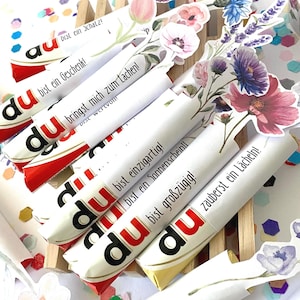 Pu&ograve; includere: Barrette di cioccolato confezionate con decorazioni floreali. Ogni barretta presenta la parola "du" e una frase in tedesco. Le confezioni sono decorate con vari fiori. Coriandoli multicolori circondano le barrette.