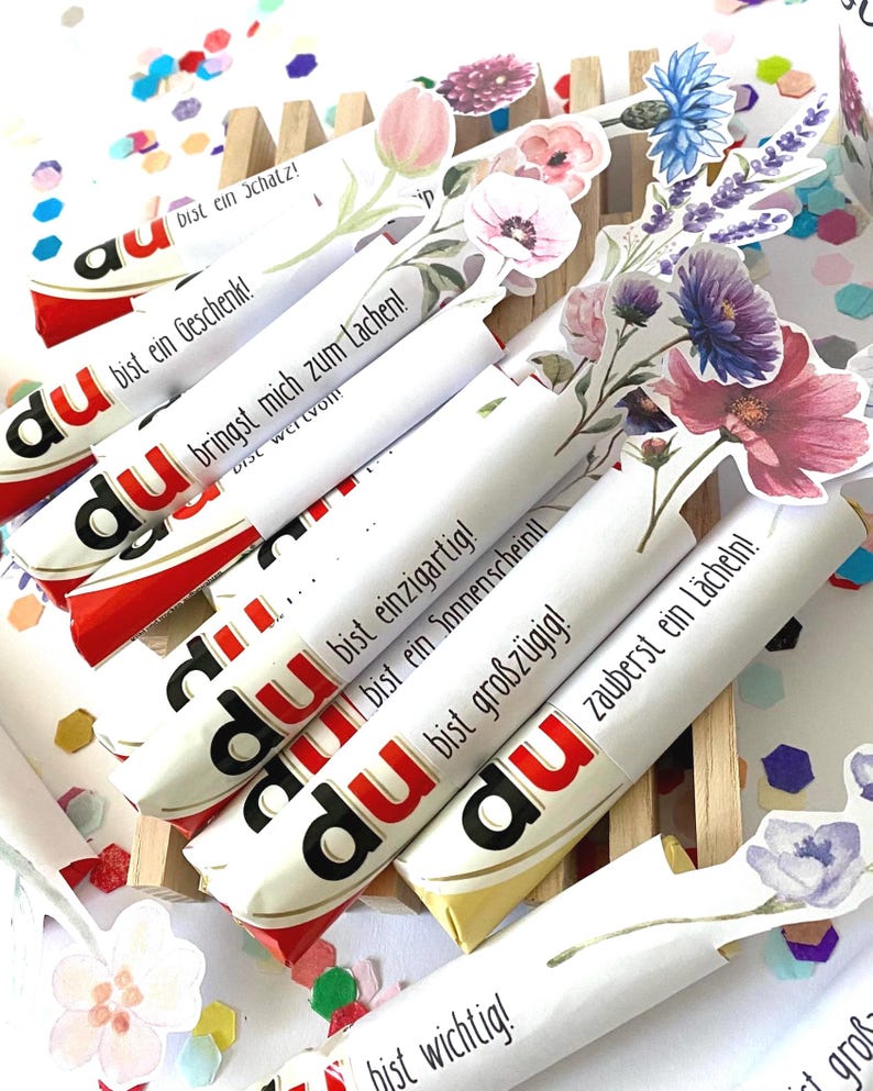 Pu&ograve; includere: Barrette di cioccolato confezionate con decorazioni floreali. Ogni barretta presenta la parola "du" e una frase in tedesco. Le confezioni sono decorate con vari fiori. Coriandoli multicolori circondano le barrette.