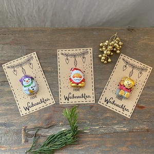 Puede incluir: Tres chocolates navideños en tarjetas decorativas. Las tarjetas presentan un muñeco de nieve, Papá Noel y un oso, cada uno con las palabras "Frohe Weihnachten". Las tarjetas están sobre una superficie de madera con una ramita de vegetación y adornos dorados.