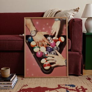May include: A framed print featuring a pink background with hands reaching towards a triangle rack of billiard balls. The balls are a mix of solid colors and disco ball designs. The hands wear watches and rings.