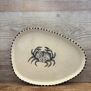 May include: An irregular, off-white ceramic plate featuring a detailed black line drawing of a crab in the centre. The plate has a speckled finish and a decorative rim with a repeating pattern of short, vertical black dashes along the edge.