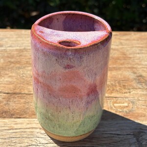 May include: A cylindrical ceramic vessel featuring a smooth, glazed finish with a gradient of colours transitioning from a light green base to a soft pink and purple top. The rim includes a distinct notched opening with a small circular hole.