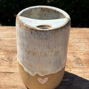 May include: A cylindrical ceramic tumbler with a speckled tan base and a smooth, off-white glazed top section. The vessel features a small, light pink heart detail on the lower portion and a circular opening on the top rim edge.