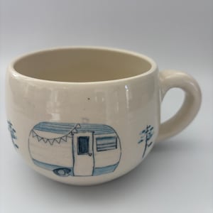 May include: A cream-coloured ceramic mug featuring a hand-drawn blue illustration of a vintage camper trailer with bunting flags above the door. The design includes small blue pine trees on either side of the camper, set against a smooth, glossy finish.