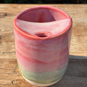 May include: A cylindrical ceramic vessel featuring a glossy glaze that transitions from a vibrant pink at the top to a soft sage green near the base. The top edge includes a curved, integrated shelf with a small circular opening in the centre.