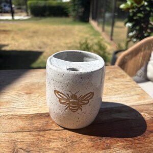 May include: A speckled, light grey ceramic toothbrush holder with a gold-coloured bee design. The holder has a rounded shape with a single opening at the top. The bee is centered on the front. The holder is on a wooden surface.
