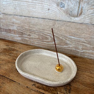May include: A speckled, oval ceramic incense holder in a neutral tone. A gold-coloured incense stick holder is in the centre, holding a brown incense stick. The holder sits on a wooden surface.