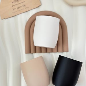 Könnte beinhalten: Eine Sammlung von drei modernen Pflanzgefäßen mit geschwungenen Seiten in Weiß, Beige und Schwarz. Ein Holzschild mit Text und ein braunes, bogenförmiges Dekorationselement sind ebenfalls zu sehen. Die Pflanzgefäße sind auf einer weißen, strukturierten Oberfläche angeordnet.