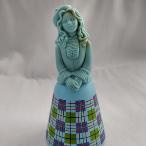 May include: A light blue ceramic figurine of a woman in a long dress with a plaid pattern. The woman has long curly hair and is wearing a hat. The dress has a blue, green, red, and black plaid pattern.