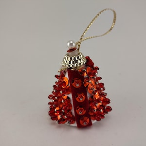 May include: A festive Christmas ornament shaped like a bell. The ornament features red sequins and beads, with a gold-colored top and hanging loop. A white base and red velvet stripe add to the design.