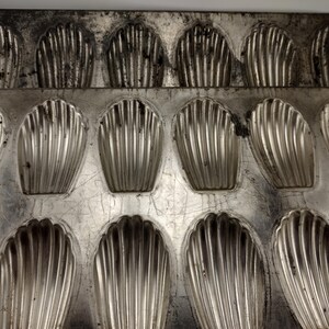 May include: A well-used, vintage madeleine pan with 12 shell-shaped molds. The aluminum pan shows signs of wear and tear, with scratches and discoloration. The molds are ribbed, designed for baking the classic French cookie.