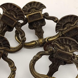 May include: Close-up of several antique bronze drawer pulls. Each pull features a decorative fan-shaped top with a rectangular plate and a curved handle. The pulls have an aged, vintage appearance, suggesting they are for furniture restoration.