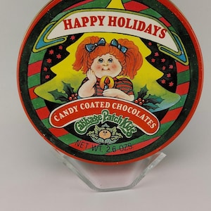 May include: Round tin of Cabbage Patch Kids candy-coated chocolates. The lid features a red-haired doll with blue bows, holding a candle, and the words "Happy Holidays." Net weight is 2.6 oz.