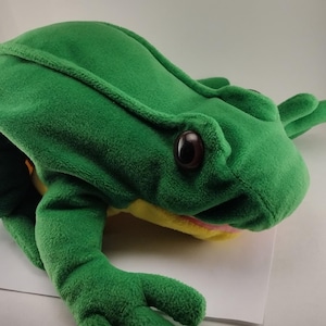 May include: A plush green frog puppet with large brown eyes, a yellow mouth, and green limbs. The frog puppet is made of soft, textured fabric and is designed to fit over a hand for play.