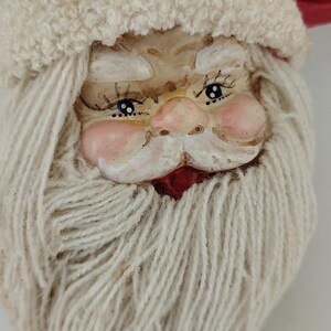 May include: A Santa Claus ornament featuring a sculpted face with rosy cheeks, painted blue eyes, and long eyelashes. The figure wears a red fabric hat with a textured white trim and has a thick, full beard made of off-white yarn with a gold string.