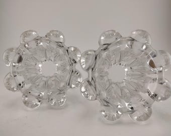 May include: Two clear, glass candle holders. Each holder has a circular design with a central opening and rounded, petal-like extensions. The glass is transparent, allowing light to pass through, and the design is symmetrical.
