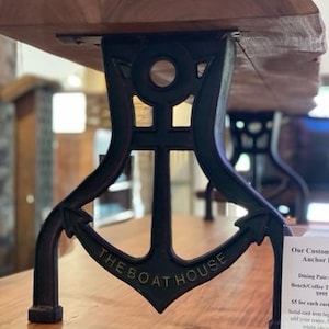 May include: Black metal anchor-shaped table legs with the words "The Boat House" on the front. The legs are supporting a wooden table top.