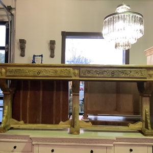 May include: A long, wooden desk with intricate carvings and two drawers. The desk has a light brown finish and is supported by four legs. The desk is sitting on top of a white dresser.