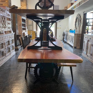 May include: A large wooden dining table with a live edge top and black metal industrial style base. The table is made of reclaimed wood and has a rustic look. The base is made of heavy-duty metal and has a gear design.