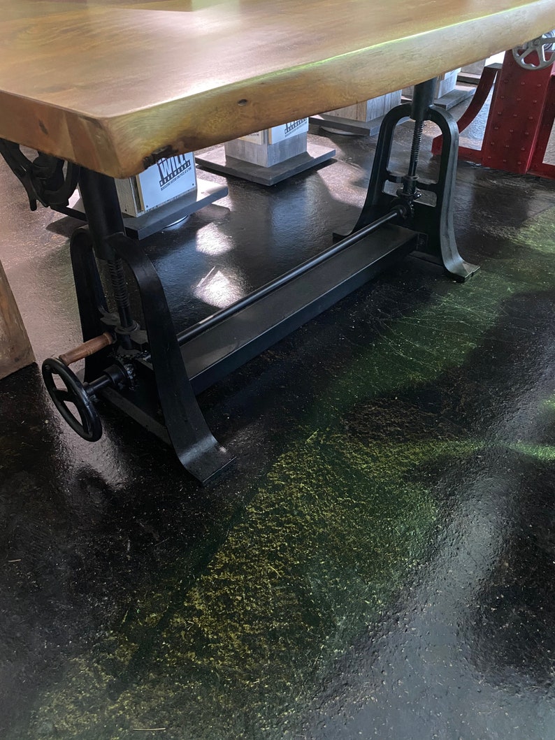 May include: A wooden table top with a live edge sits on a black metal adjustable base with a large hand crank. The base has a long metal bar that runs the length of the table. The table is on a black floor with green and yellow flecks.