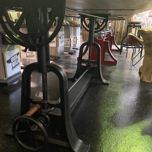 May include: A black metal table base with a crank and a wooden handle. The base is adjustable in height and has a long metal bar running along the bottom. The table base is in a showroom setting.