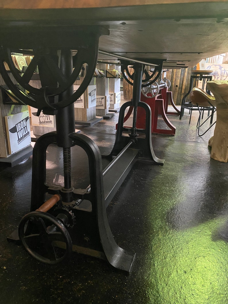 May include: A black metal table base with a crank and a wooden handle. The base is adjustable in height and has a long metal bar running along the bottom. The table base is in a showroom setting.