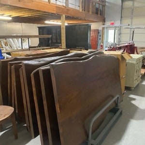 May include: A stack of large, dark brown wooden slabs with a live edge. The slabs are stacked on top of each other and are leaning against a wall. The slabs are likely for use as tabletops or countertops.