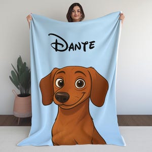 May include: A light blue blanket with a cartoon illustration of a dachshund dog. The dog is brown with big eyes and a smiling expression. The name "DANTE" is written in black script. The blanket is displayed against a white backdrop.