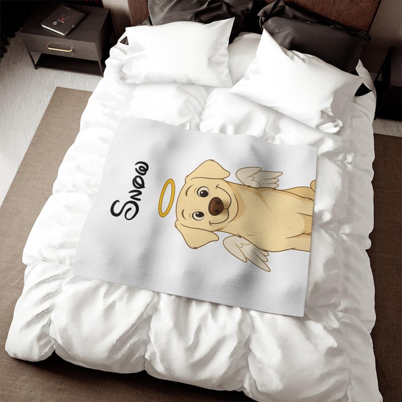 May include: A white blanket with a cartoon Labrador illustration, complete with angel wings and a halo. The word "Snow" is written in black script. The blanket is placed on a bed with white bedding.