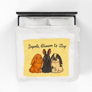 May include: A yellow blanket with three cartoon rabbits. The rabbits are orange, black, and black and white. The blanket has the names "Japmie, Graham & Lilly" written in cursive.