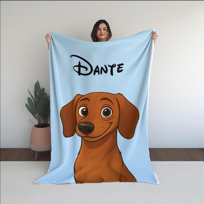 May include: A light blue blanket with a cartoon illustration of a dachshund dog. The dog is brown with big eyes and a smiling expression. The name "DANTE" is written in black script. The blanket is displayed against a white backdrop.