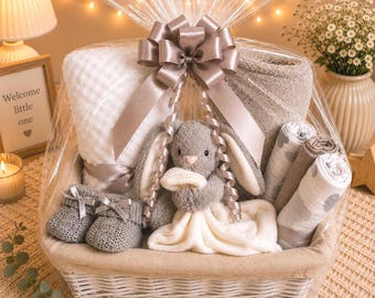 Può includere: Un cesto regalo avvolto in cellophane trasparente con un fiocco di raso, contenente una coperta bianca a nido d'ape, una coperta in peluche grigia, un doudou a forma di coniglio grigio, scarpine in maglia grigie e tre teli avvolti.