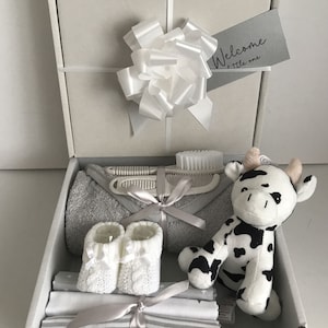 May include: A white gift box with a white bow and a tag that says "Welcome little one". The box is filled with baby items, including a grey towel, white booties, a stuffed cow toy, and a set of baby grooming tools.