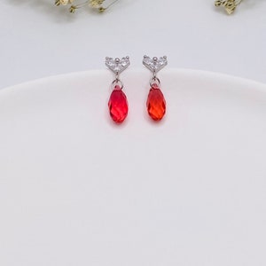 May include: A pair of silver earrings with a small heart-shaped design and a dangling red teardrop-shaped crystal.