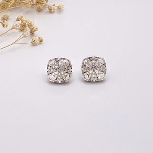 May include: A pair of cushion-cut stud earrings featuring clear, faceted stones that reflect light with a brilliant sparkle. The stones are set in a simple, polished metal frame, showcasing a square shape with rounded corners and a classic design.