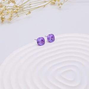 May include: A pair of small, square-shaped earrings with a light purple crystal in the centre. The earrings are on a white background.