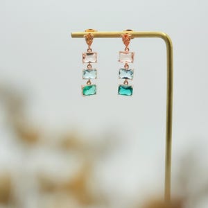 May include: A pair of dangle earrings featuring three rectangular gemstones in shades of pink, blue, and green. The earrings are made of gold-toned metal.