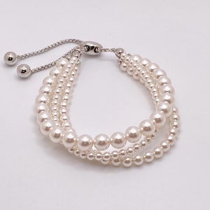 May include: A multi-strand bracelet featuring white pearls of varying sizes arranged in three rows. The bracelet includes a silver-toned adjustable sliding clasp mechanism with two polished metal spheres at the ends of the chain links.